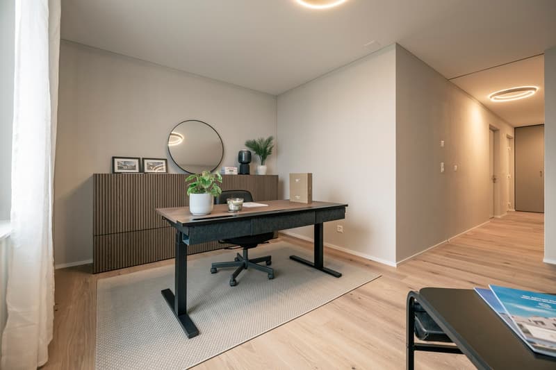 The image shows a modern and minimalist home office space. The room features a large adjustable desk with a wooden top and a black metal frame. There is a round mirror on the wall above the desk, and a cabinet or credenza with vertical slats along the wal