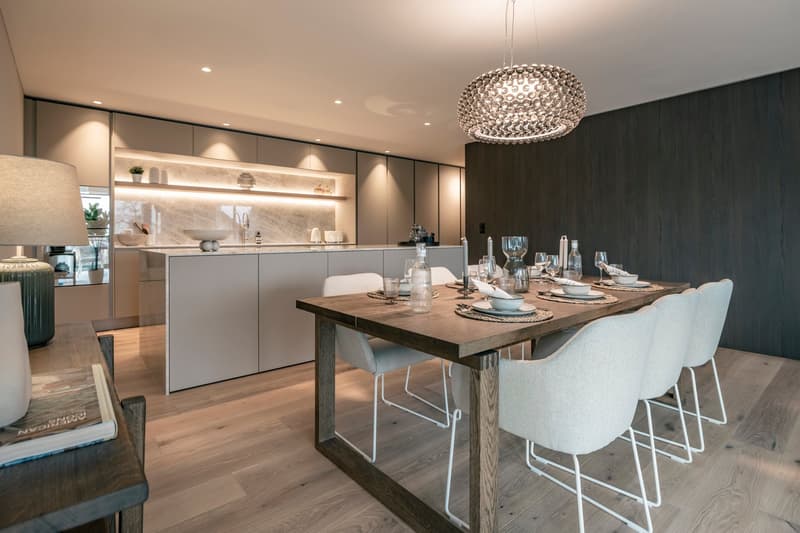 The image shows a modern and stylish kitchen and dining area. The kitchen features gray cabinets, a marble backsplash, and built-in shelving. The dining table is made of wood with a metal frame, and is surrounded by white upholstered chairs. A large, crys