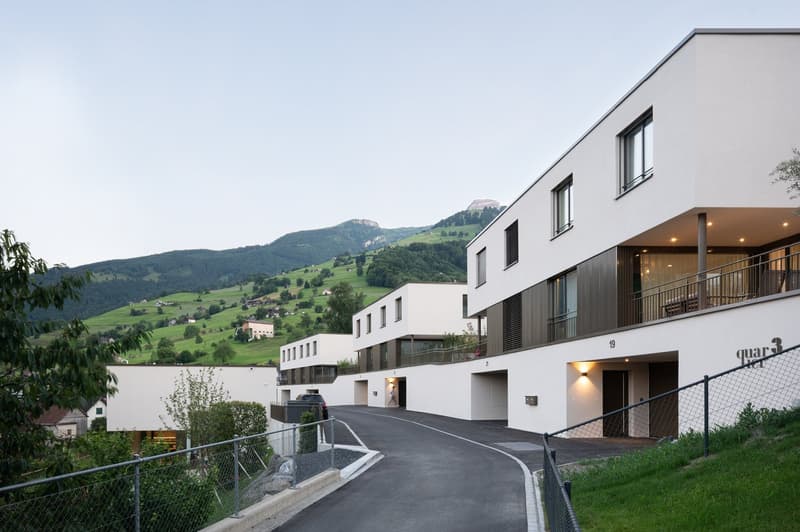 A modern, multi-story apartment building with balconies overlooking a scenic mountainous landscape. The building has a clean, minimalist design with large windows and a paved driveway leading to the entrance.