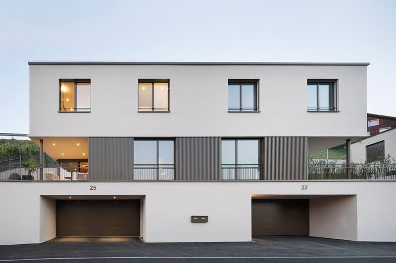2-story modern apartment building with a flat roof, gray exterior, and large windows. The building has two garage doors on the ground floor, indicating the presence of a garage. There are also two parking spaces visible in front of the building.
