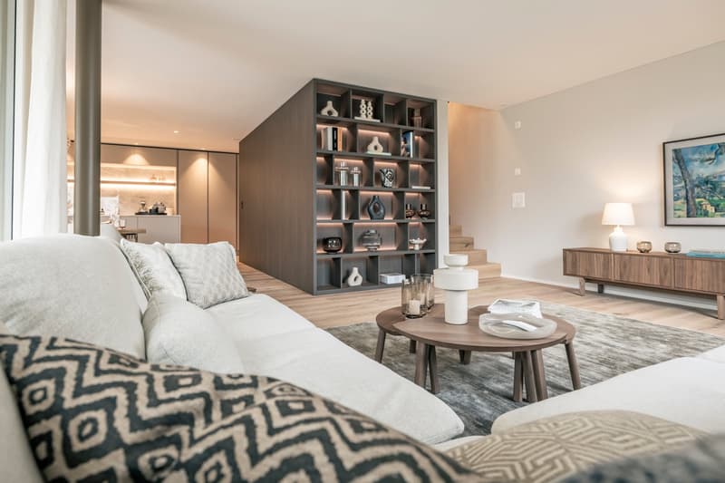 Spacious living room with a large gray sectional sofa, a wooden coffee table, and a built-in shelving unit displaying decorative items. The room has hardwood floors, neutral-colored walls, and a fireplace visible in the background.
