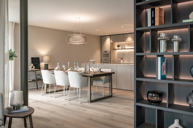 Spacious open-plan kitchen and dining area with modern, minimalist design. The kitchen features sleek gray cabinets, built-in appliances, and a marble backsplash. The dining table is made of wood with a metal frame, surrounded by white upholstered chairs.