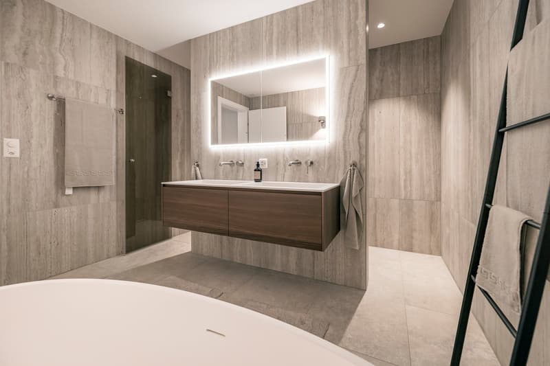 Large bathroom with gray marble-like tiles on the walls and floor, a floating vanity with double sinks, a large illuminated mirror, and a glass shower enclosure.