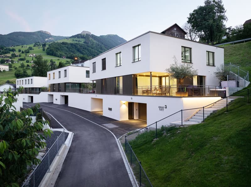 Modern, multi-story building with a white exterior and large windows. It has a balcony or terrace on the upper level. The building is situated in a scenic, mountainous landscape with green hills and trees in the background.