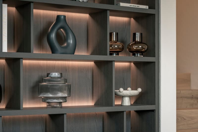 Wooden shelving unit with various decorative items, including a black vase, two glass vases, a metal sculpture, and a ceramic bowl. The shelves are illuminated, creating a warm and cozy atmosphere.
