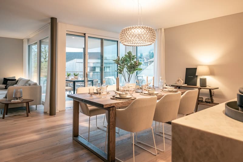 Spacious dining area with large windows overlooking a balcony, wooden dining table with chairs, a decorative chandelier, and a living room visible in the background.