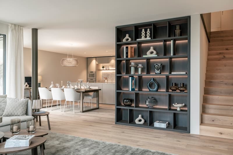 The image shows an open concept living room and kitchen area. The living room has a large built-in shelving unit with decorative items displayed, and a fireplace is visible. The kitchen area has modern appliances and cabinetry, and a dining table with whi