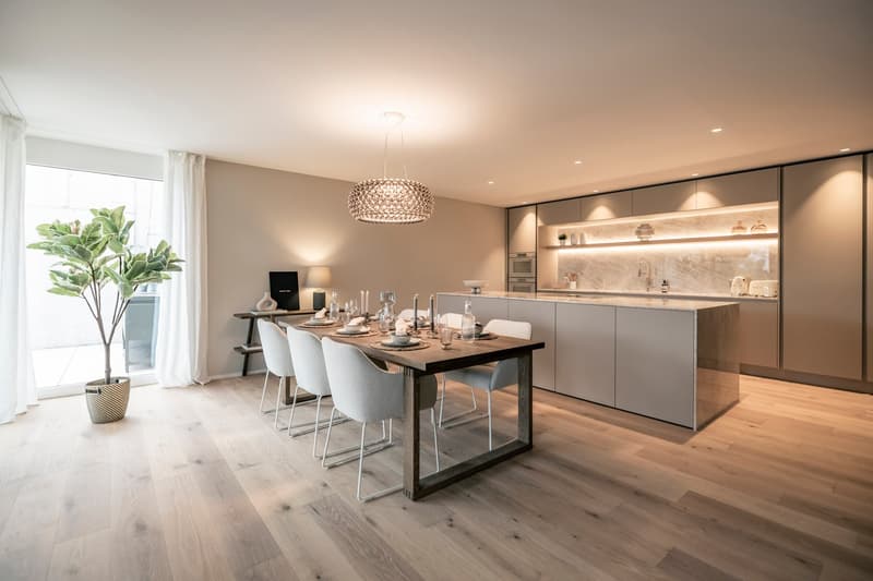 Spacious open-plan kitchen and dining area with modern, minimalist design. The kitchen features sleek gray cabinets, a marble backsplash, and built-in appliances including a dishwasher. The dining table is made of wood with a metal frame, surrounded by gr