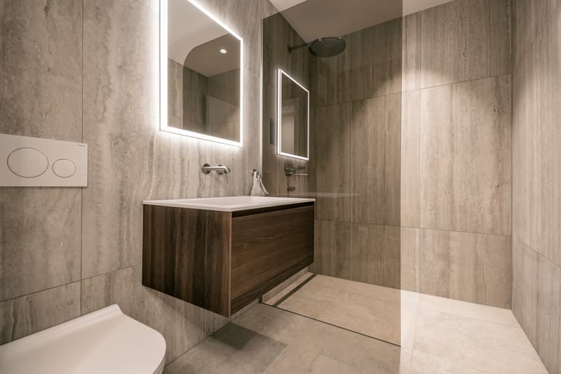 Modern bathroom with gray marble-like tiles, a wooden vanity unit with a sink, a large mirror with LED lighting, and a shower area with a rain shower head.