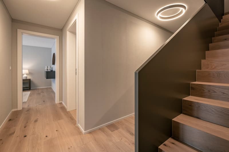 The image shows a modern and minimalist hallway with hardwood floors, white walls, and a circular lighting fixture on the ceiling. The hallway leads to another room, which can be seen through the open doorway. The stairs on the right side of the hallway h