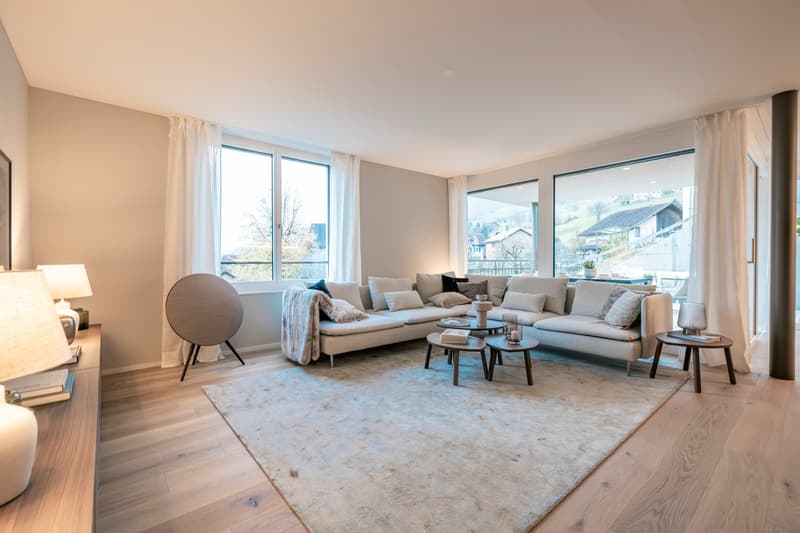 Spacious living room with large windows offering a scenic view, comfortable L-shaped sofa, coffee table, and side tables. The room has a neutral color scheme with light hardwood floors and white walls.