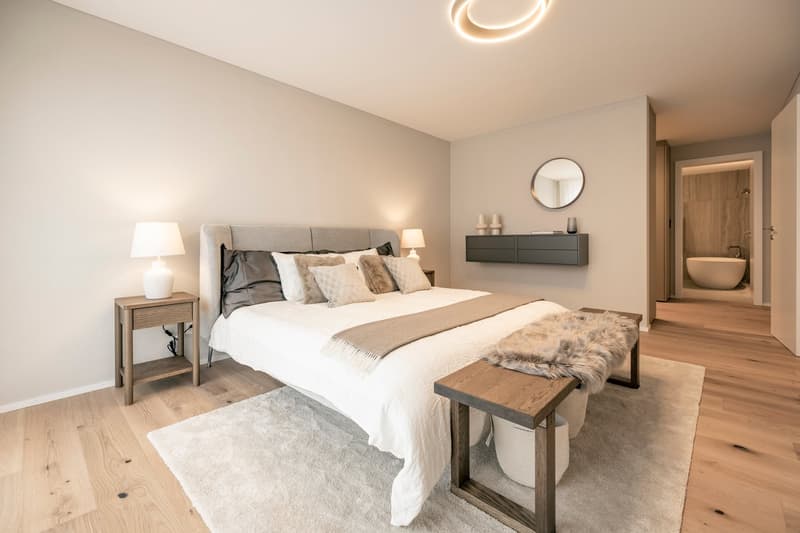 Spacious bedroom with a large bed, nightstands, a dresser, and a round mirror on the wall. The room has hardwood floors and a cozy rug. There is also a bench at the foot of the bed and a doorway leading to an ensuite bathroom.