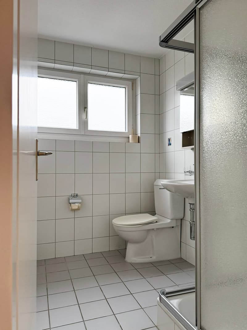Small white tiled bathroom with toilet, sink, and glass shower door
