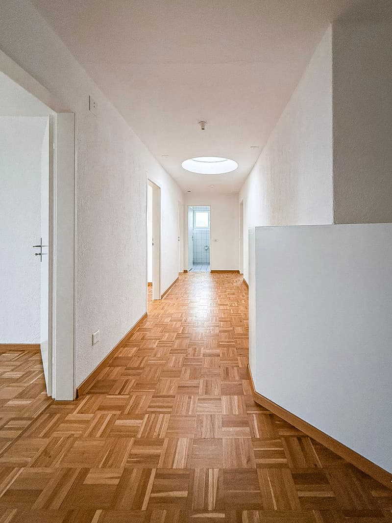Empty hallway with wooden floors, white walls, sky light, power outlet, entrance doors, and an exit door leading to a room.