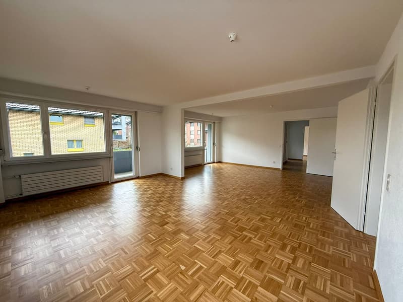 Open room with wooden parquet flooring, glass windows, glass balcony doors, and a radiator against the wall.