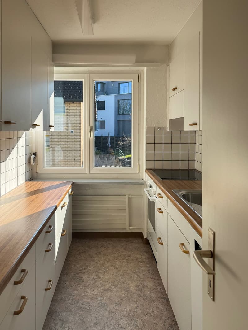 A kitchen with a wooden countertop, white cabinets, gas stove, and a sink with a faucet. There is a window with a view of the outside. The room has white walls, tiled backsplash, and a white ceiling. A radiator is installed on the wall near the window. A