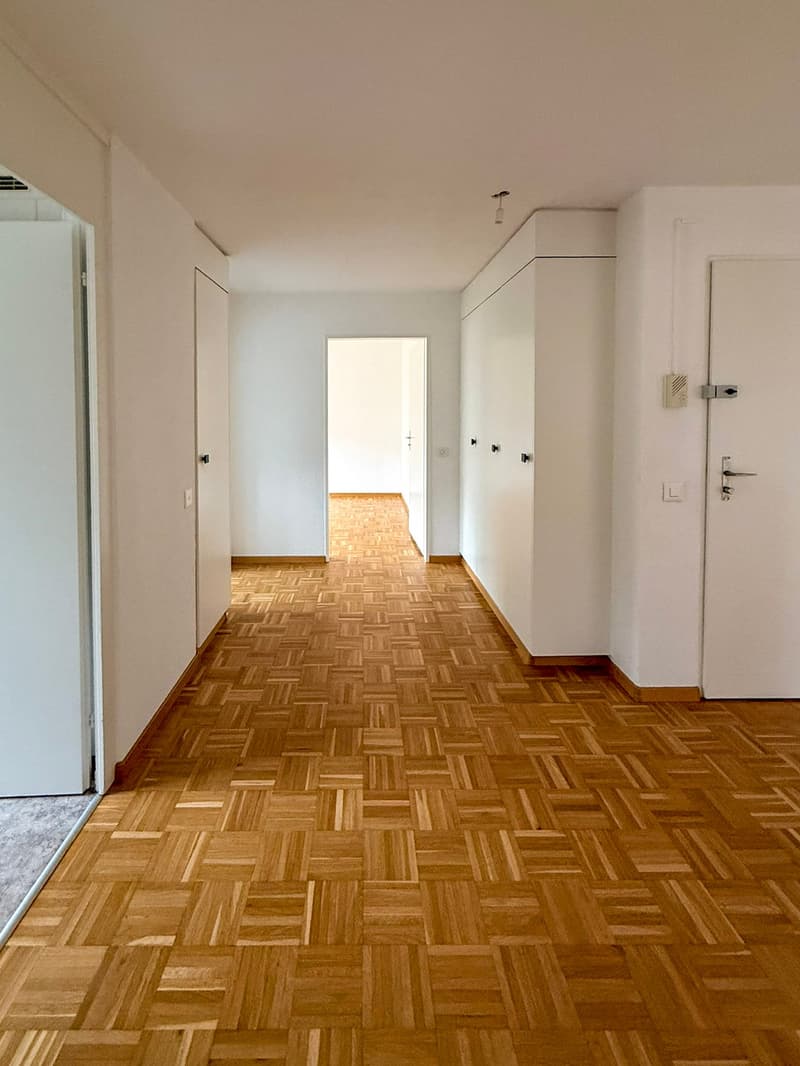 Empty hallway with wooden parquet flooring, white walls, two doors, and one open door