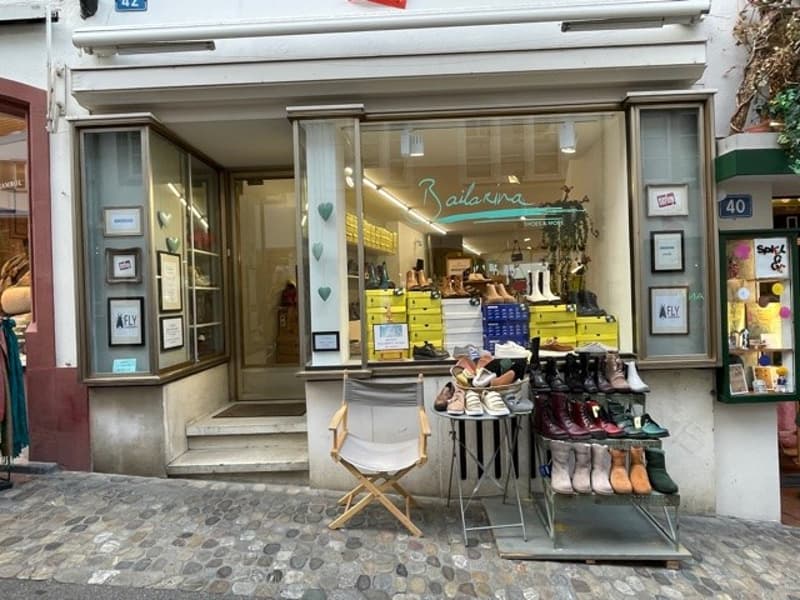 Storefront with display windows showcasing shoes, wooden chair in front, shelves with boots, white facade, steps leading up to entrance.