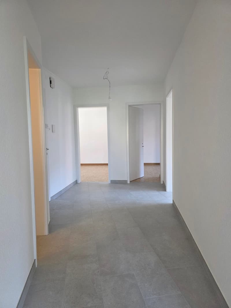 Empty hallway with tiled floor, three doors, one leading to a room with wooden floor