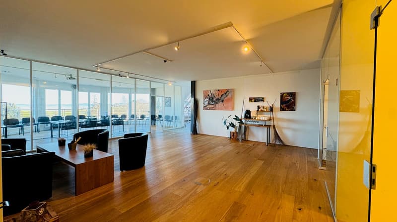 open room with large glass walls, wooden floor, view of trees, empty chairs and tables, a table, a plant, and a painting