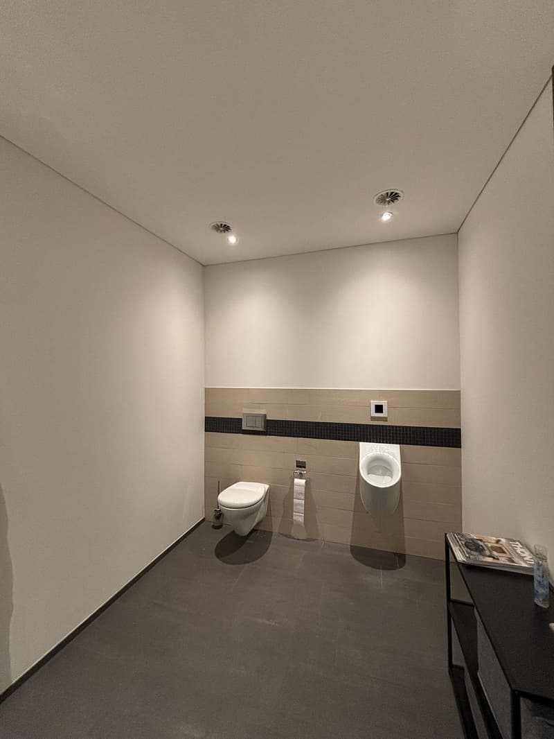 modern design, white toilet, urinal, tiled floor, white walls, towel holder, black shelf with magazine