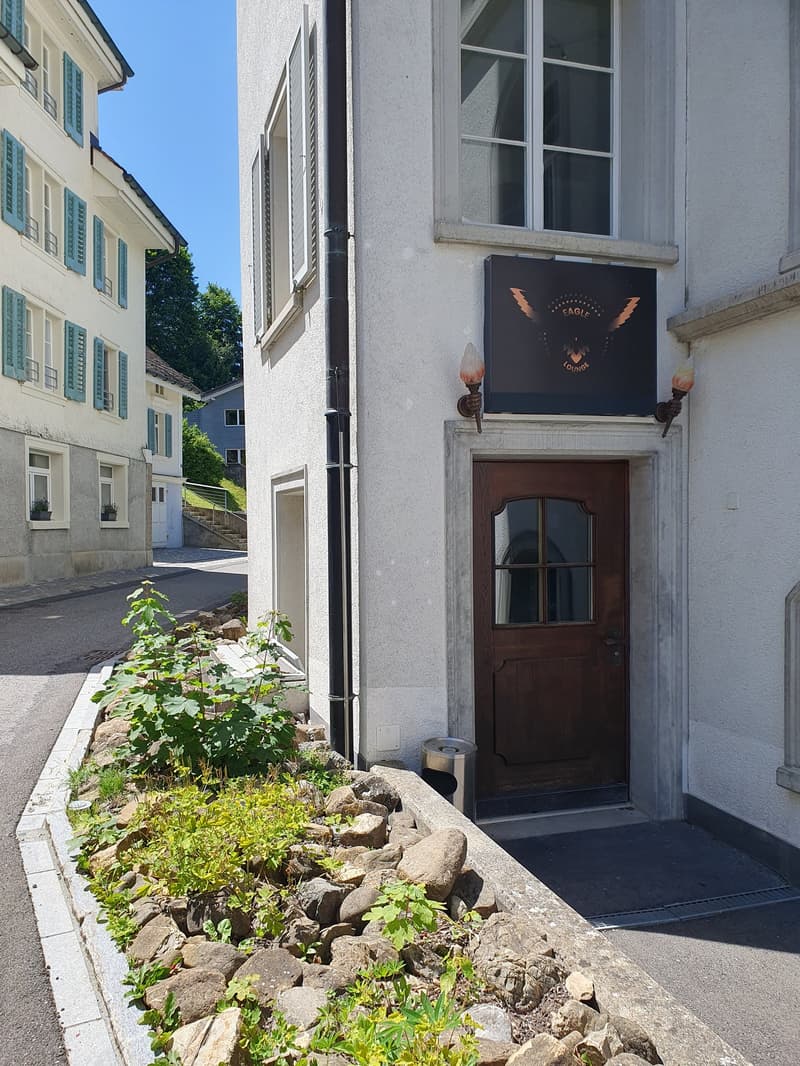 White building with brown door, black sign above door, lamp on either side of sign, small garden in front, rocks and plants