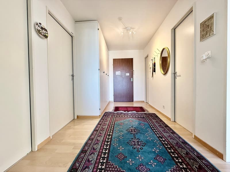 Empty hallway with wooden floors, white walls, two rugs, a mirror, and a ceiling fan.