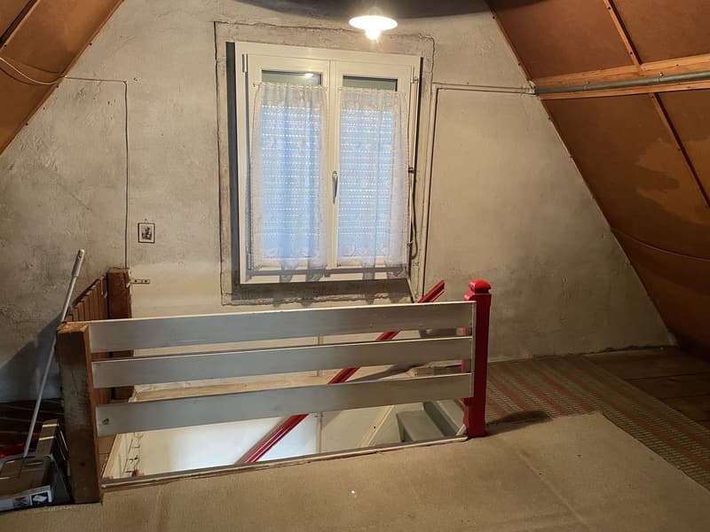 a small room with a window and a ceiling-hung light fixture, wooden handrail on stairs, wooden floor, wall-mounted picture frame, white window with sheer curtains