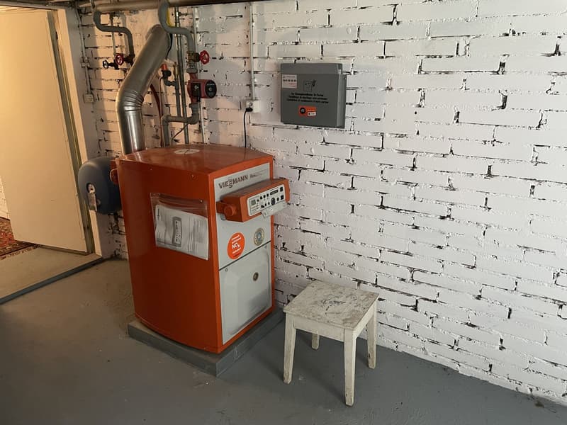 Room with a white brick wall, an orange gas heater, a gray electrical box, a small white stool, and an open door with a carpet