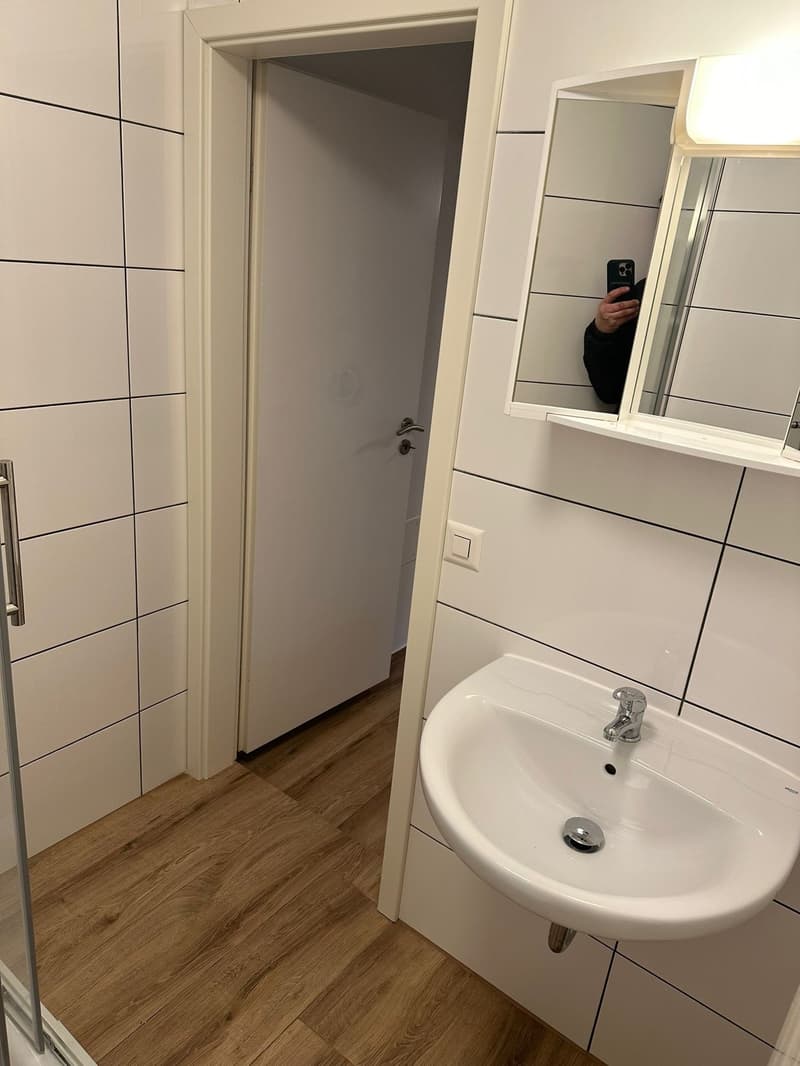 Bathroom with white tiles, wooden floor, white sink, mirror, and shower enclosure