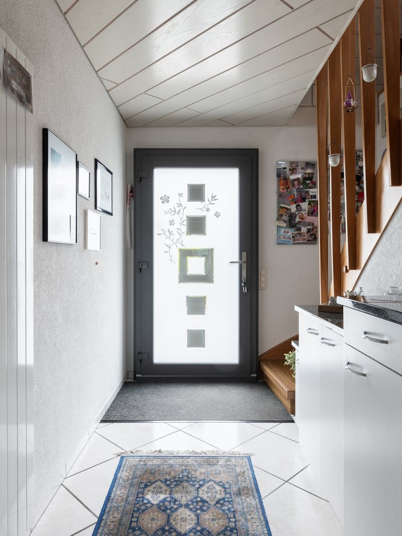 The image shows an entryway with a patterned tile floor, a wooden bench, and a decorative rug. The walls are white with framed artwork, and the ceiling has a diagonal wood paneling design. The main feature is the glass-paneled front door with floral desig