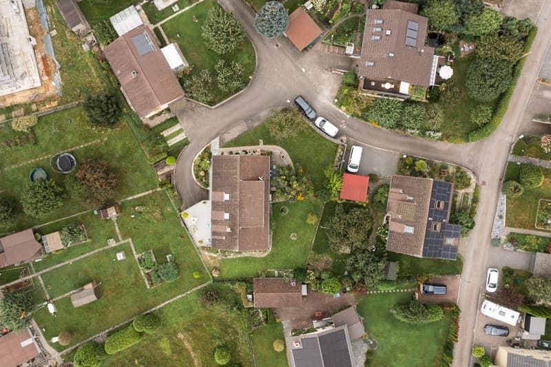 The image shows an aerial view of a residential area with several houses and buildings surrounded by lush greenery. The houses have red tile roofs and there are paved roads and driveways with parked cars visible. The area appears to be a well-maintained a
