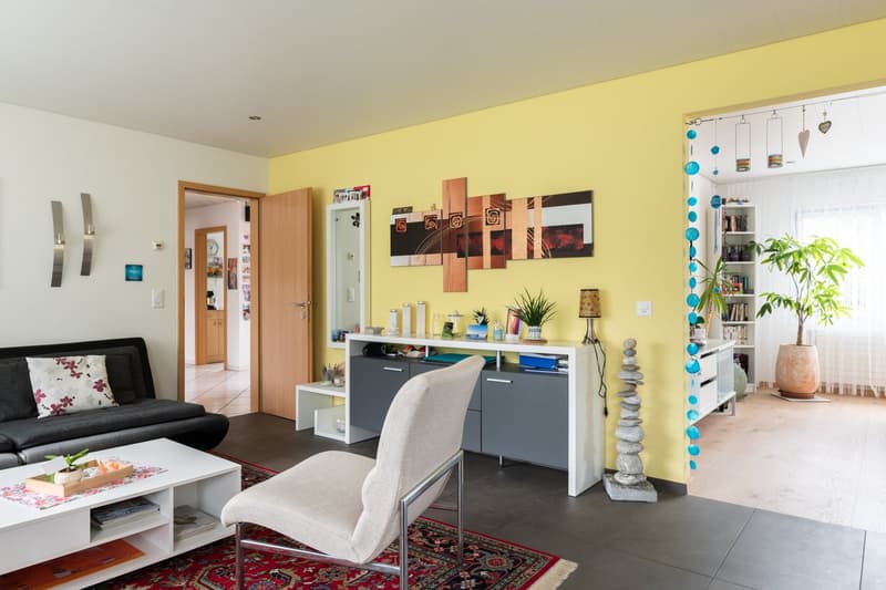 Bright and colorful living room with yellow walls, modern furniture, artwork, and decorative elements. The room features a sofa, coffee table, shelving unit, and a desk with a chair. There is also a balcony or terrace visible, and a dishwasher can be seen