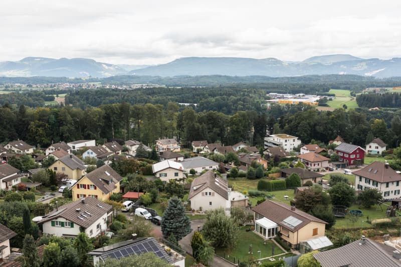 Aerial view of a residential area surrounded by mountains and forests. The image shows a village with various houses, some with balconies or terraces, and parking spaces visible. The landscape is characterized by rolling hills, dense forests, and distant