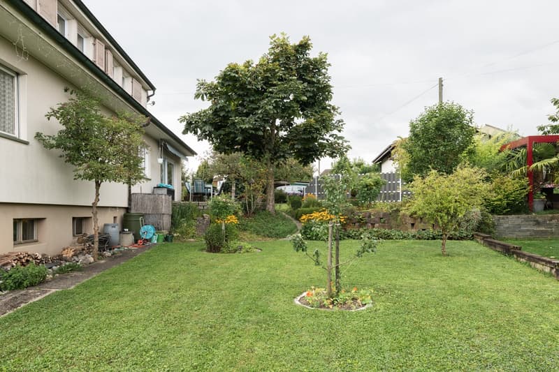 Two-story residential building with a well-maintained garden, featuring a lush lawn, mature trees, and various flowering plants. The building has a balcony or terrace, and there appears to be a parking area on the property.