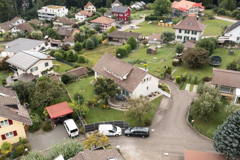 This image shows an aerial view of a residential neighborhood with a mix of single-family homes and multi-unit buildings. The houses have red tile roofs and are surrounded by lush greenery, including trees and gardens. There is a paved driveway or parking
