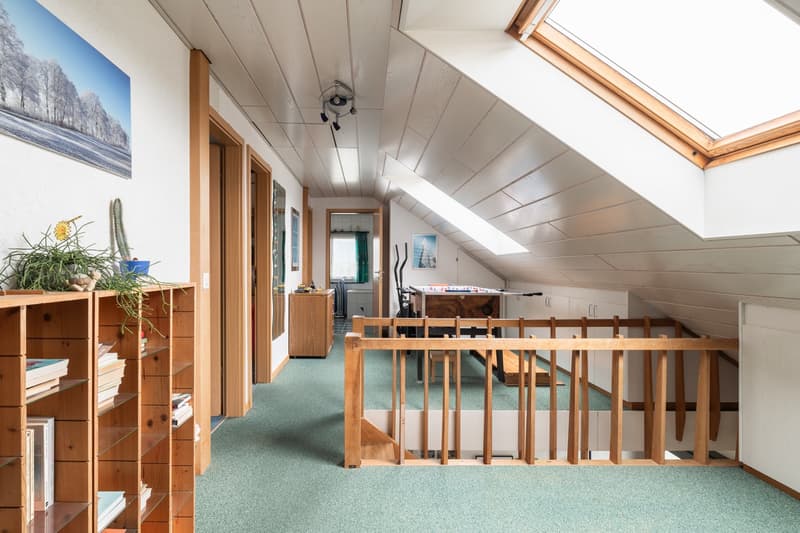 Spacious attic or loft space with wooden beams, slanted ceiling, and skylights. The room features wooden furniture, shelves, and a staircase leading to the upper level. There is also a large window or balcony door providing natural light and a view.