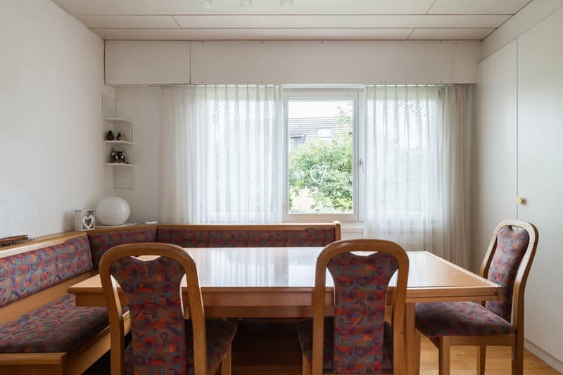 Spacious dining area with a wooden table and chairs, a built-in bench seating along the wall, and a large window overlooking the outdoor greenery.