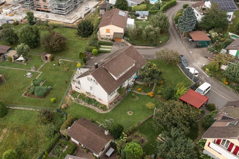 Two-story residential house with a red tile roof, surrounded by a well-maintained garden with various trees and shrubs. There is a garage and parking space visible on the property. The house appears to be located in a residential neighborhood with other h