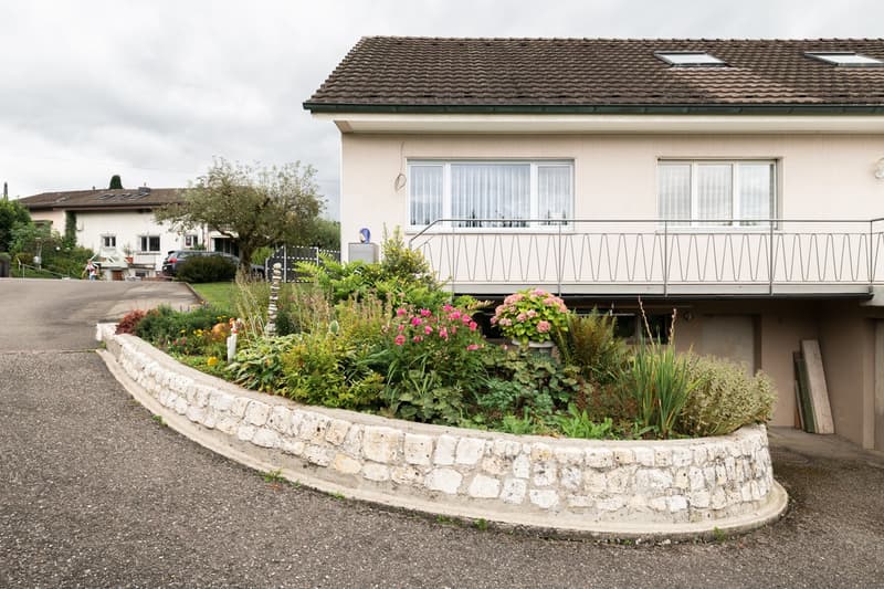 Two-story house with a tiled roof, white exterior walls, and large windows. The house has a balcony or terrace on the upper floor. There is a stone retaining wall surrounding a well-maintained garden with various flowering plants and shrubs.