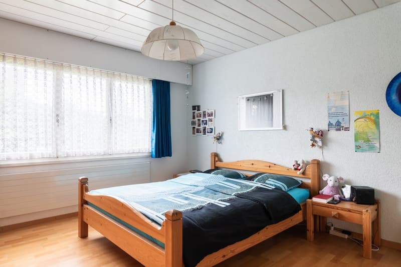 Spacious bedroom with wooden bed frame, bedside tables, curtains, and wall decor. Large windows provide natural light and a view.
