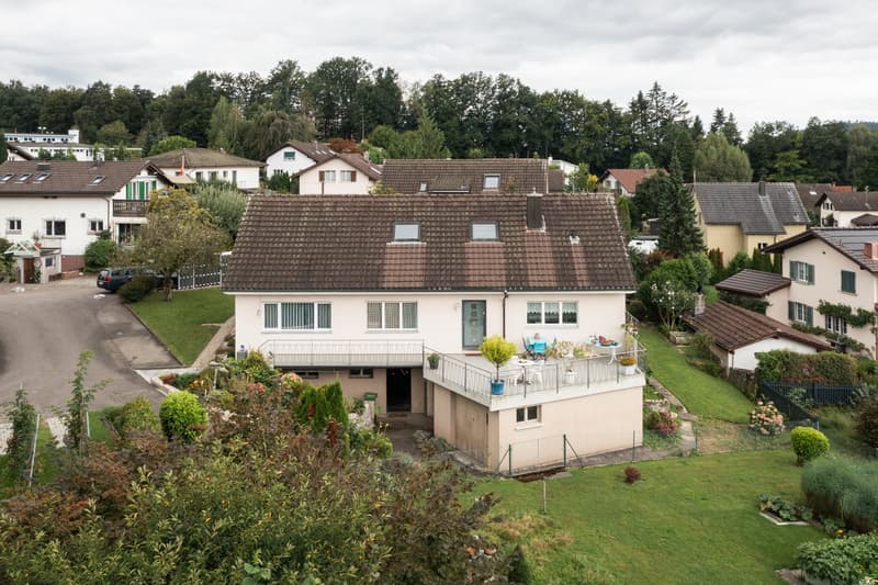 Two-story house with a brown tiled roof, white exterior walls, and a balcony on the upper level. The house is surrounded by a well-maintained garden with trees and other vegetation. There are other residential buildings visible in the background.