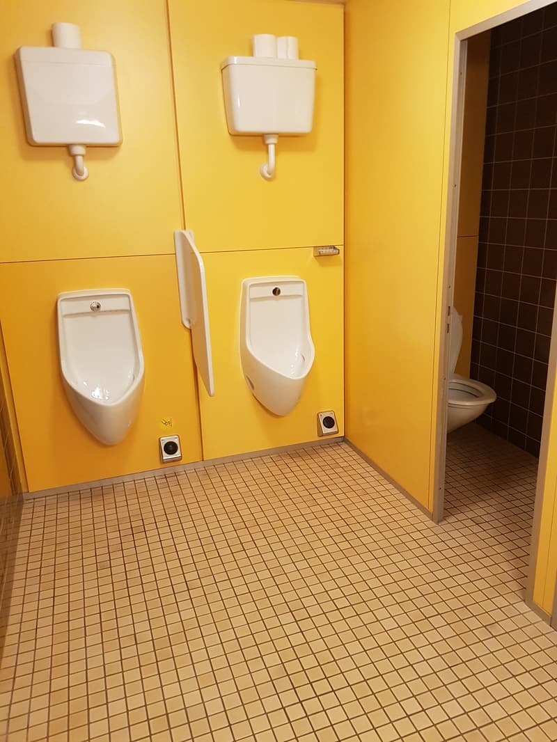 The image shows a bathroom with yellow walls, white tile flooring, a urinal, and a toilet. There are also two wall-mounted fixtures that appear to be hand dryers or soap dispensers.