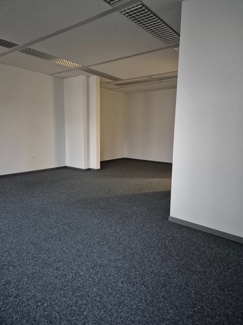 Empty room with white walls, gray carpet flooring, and white ceiling with grid vents