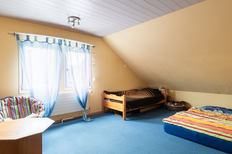 two beds, one small wooden, one on floor, window with blue curtains, wooden desk, chair, ceiling lamps