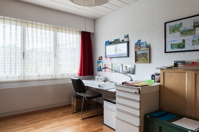 desk, lamp, office chair, framed pictures on the wall, radiator, window with curtains
