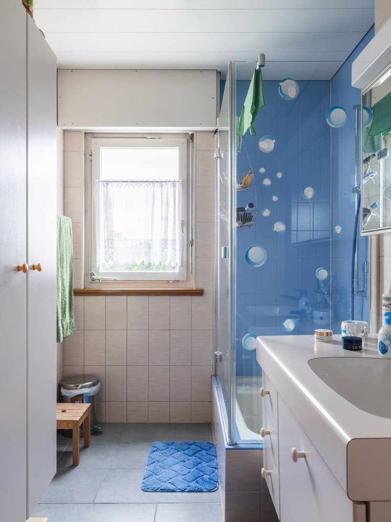White tiles, blue tile wall, window, wooden stool, trash bin, glass shower, blue rug
