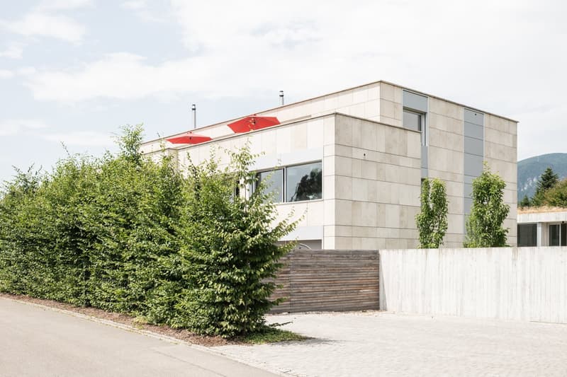 Modern house, white walls, concrete roof, two red umbrellas, balcony, hedge, wooden gate