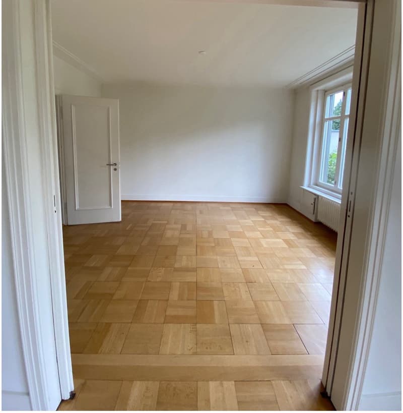 Empty room with wooden parquet flooring, white walls, white doors, window with white frame, white radiator