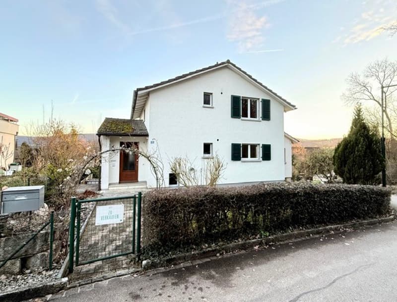 Detached house, raised ground floor, white painted exterior, brown wooden door, 5 windows, green gate, sign with text 'VERKAUFEN', greenery in front